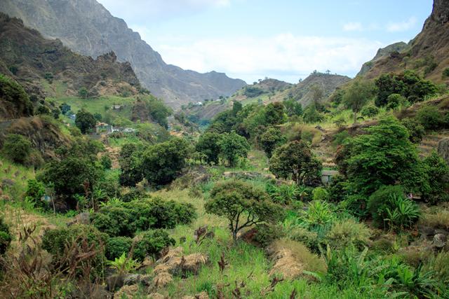 Une vallée verdoyante au Cap-Vert, entourée de montagnes rocheuses brunes, est remplie d’arbres verts et de petites maisons colorées sous un ciel partiellement nuageux.
