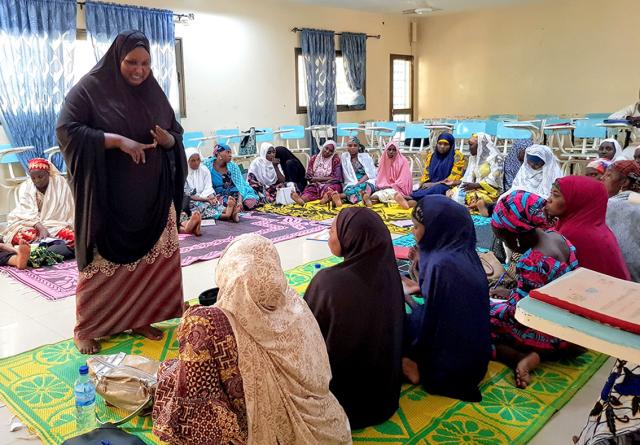 Une femme en hijab noir parle à un groupe assis sur des tapis colorés dans une salle, avec des chaises bleues et des rideaux en arrière-plan.