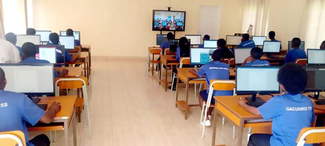 Students in blue uniforms sit at desks with computers in a Rwanda classroom, facing a TV screen.