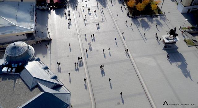 A large, sunny plaza in Kosovo is filled with people walking, casting long shadows. A statue of a rider on horseback stands on the right, near a domed building.