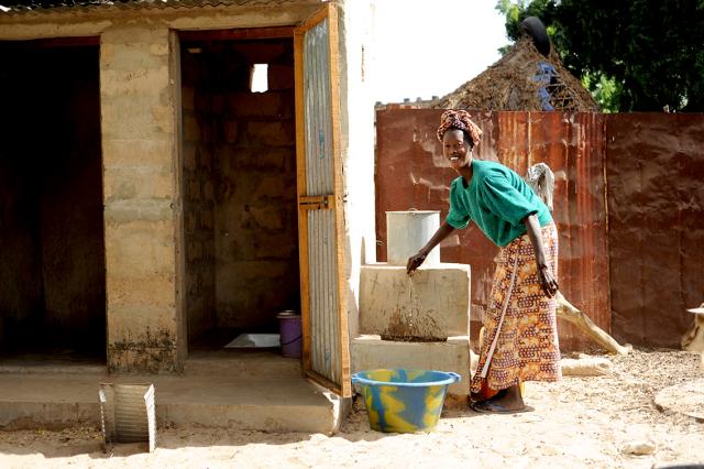 Une femme en tenue colorée lave des vêtements. Elle remplit une baassine d’eau au robinet.
