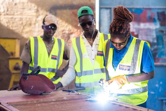 Trois apprentis ouvriers en gilets jaunes sont au premier plan. Un jeune femme soude du métal en faisant des étincelles.