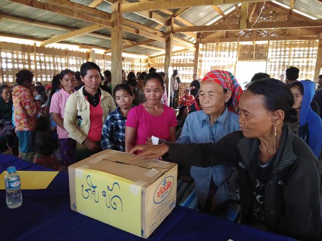 People in colorful clothing gather in a wooden structure, placing votes in a yellow box on a table.