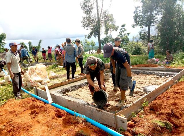 Des membres de la communauté participent au travaux du village. Deux hommes au premier plan déposent du béton afin de réaliser une dalle