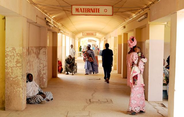 Un couloir animé de la maternité au Sénégal montre des gens marchant et assis, avec une pancarte "MATERNITÉ". Les murs sont usés, et les patients attendent en vêtements colorés.