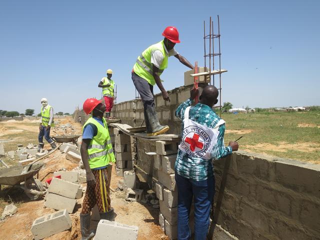 Des ouvriers en gilets fluorescents et casques construisent un mur en parpaings sur un chantier au Mali, sous un ciel bleu, avec des outils et des blocs éparpillés autour.