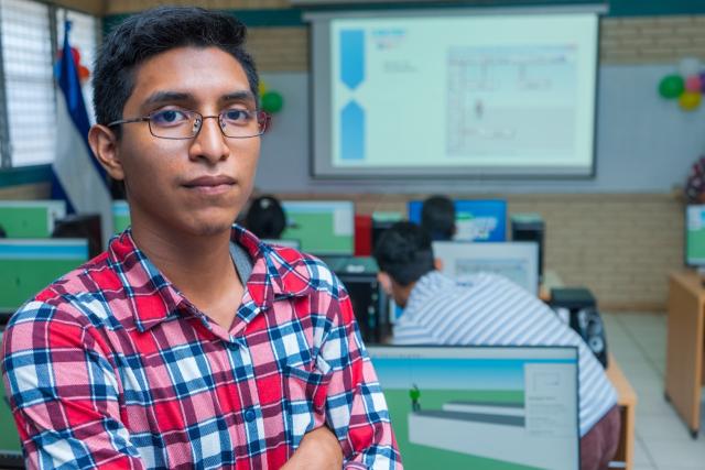 Portrait of a young Latin Central American man in glasses with his arms crossed and a slight smile standing in a classroom full of computers