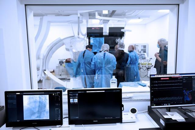 Medical staff in blue gowns perform a procedure in a sterile room, surrounded by advanced equipment. Monitors in the foreground display X-rays and vital signs.